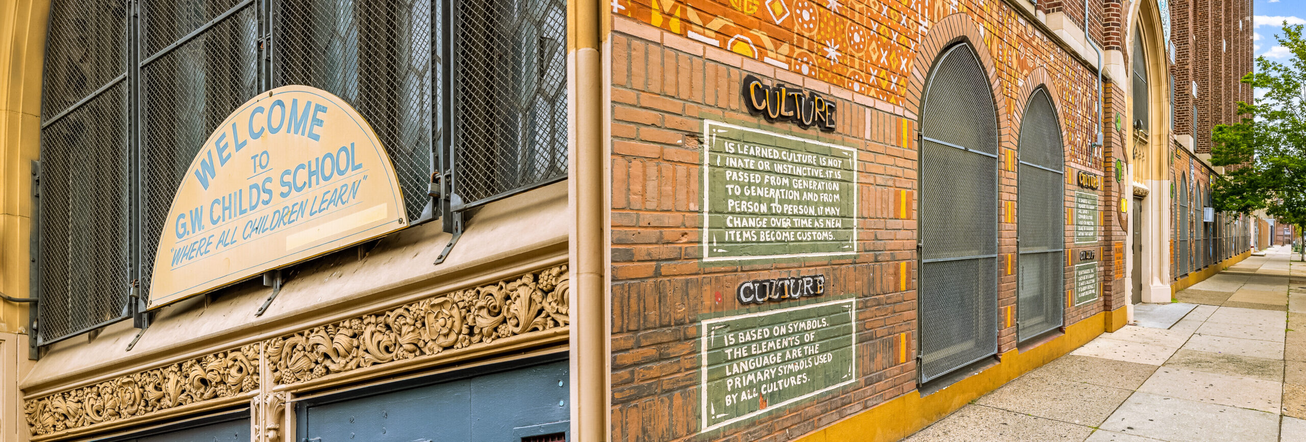 Exterior view of G.W. Childs School showing a welcome sign above the entrance and a long brick wall with colorful culture-themed murals along the sidewalk.
